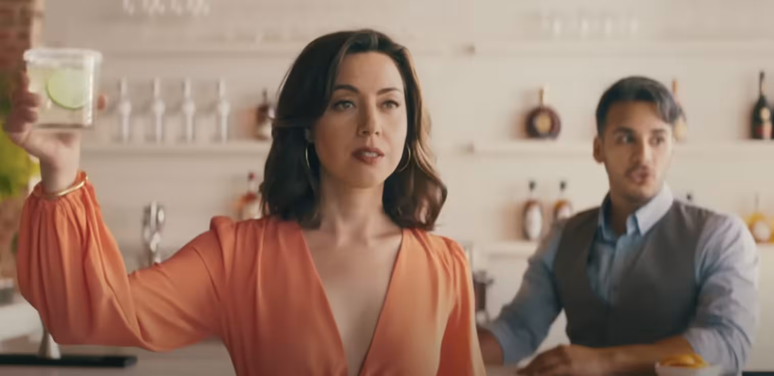 Aubrey Plaza wears a salmon blouse and holds a margarita in the air in front of a bartender