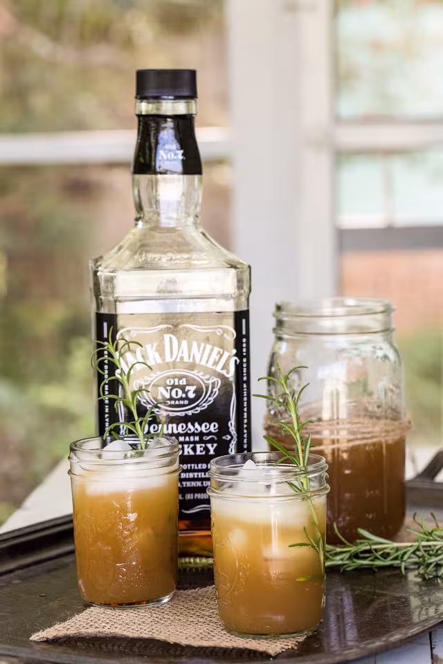 A bottle of Jack Daniels and an apple whiskey cocktail poured from a mason jar into two glasses sit on a table