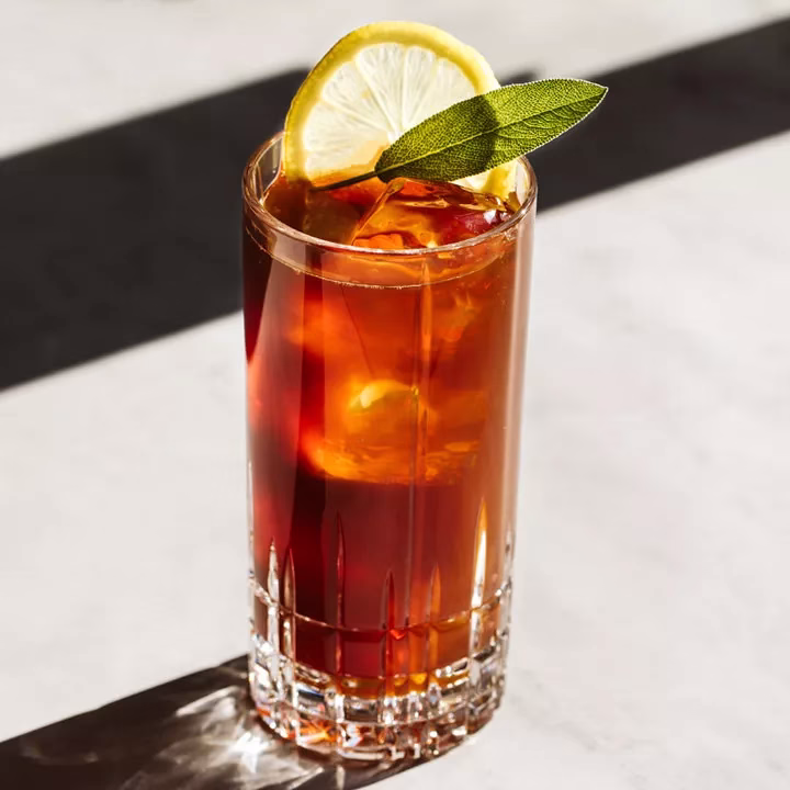 A rich brown cocktail in a Collins glass with a lemon wedge and sage leaf.